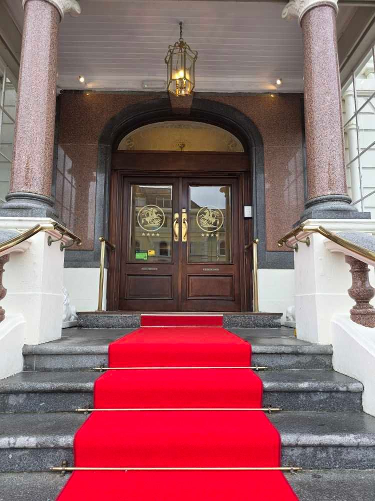 Elegant entrance with pink marble columns and a bright red carpet leading up granite stairs to wooden double doors.  St. George's Hotel Llan