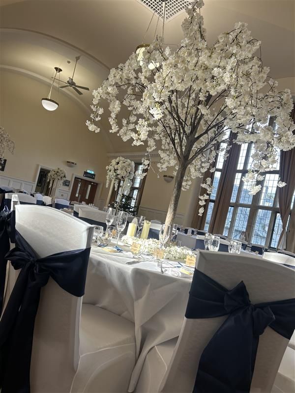 Elegant wedding reception hall with white blossom tree centerpiece, navy sash chairs, and white tablecloths in a grand venue.