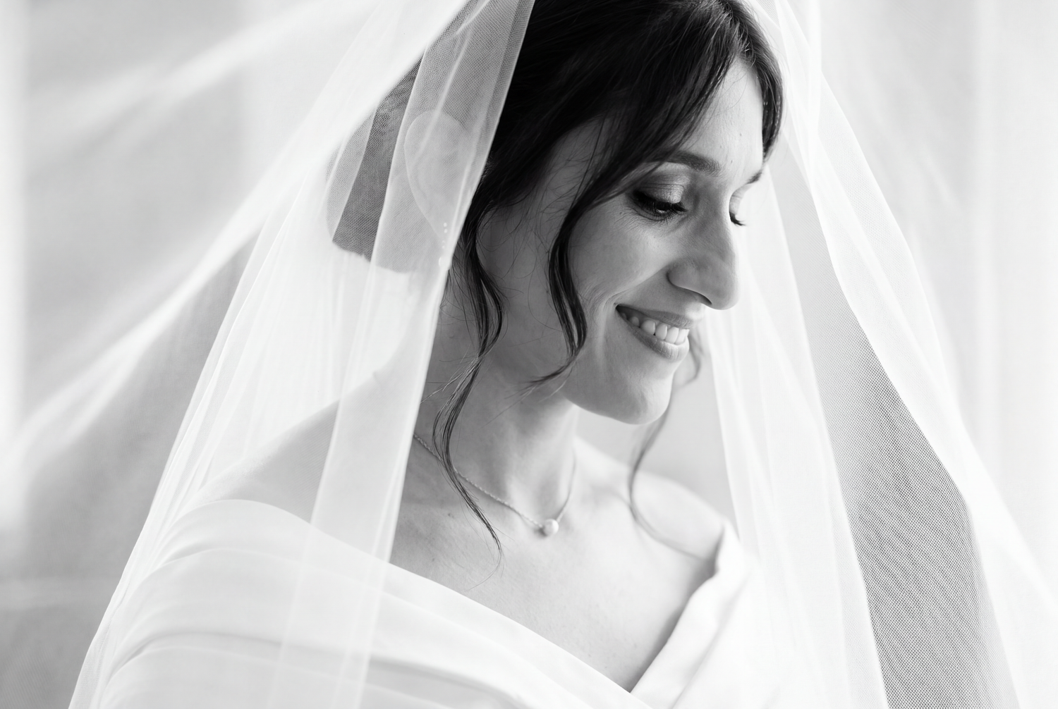 Black and white bridal portrait of smiling bride under flowing veil, wearing off-shoulder gown and delicate necklace.