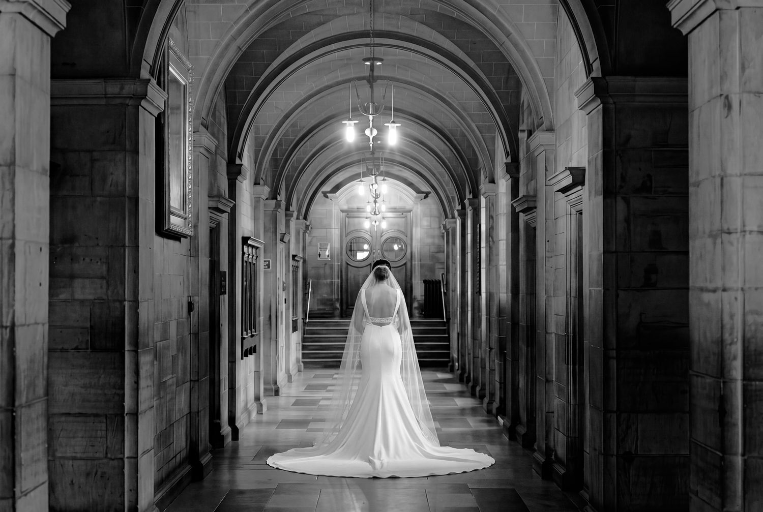 Black and white photo of a bride in a long veil walking down an elegant stone hallway with arched ceilings.
