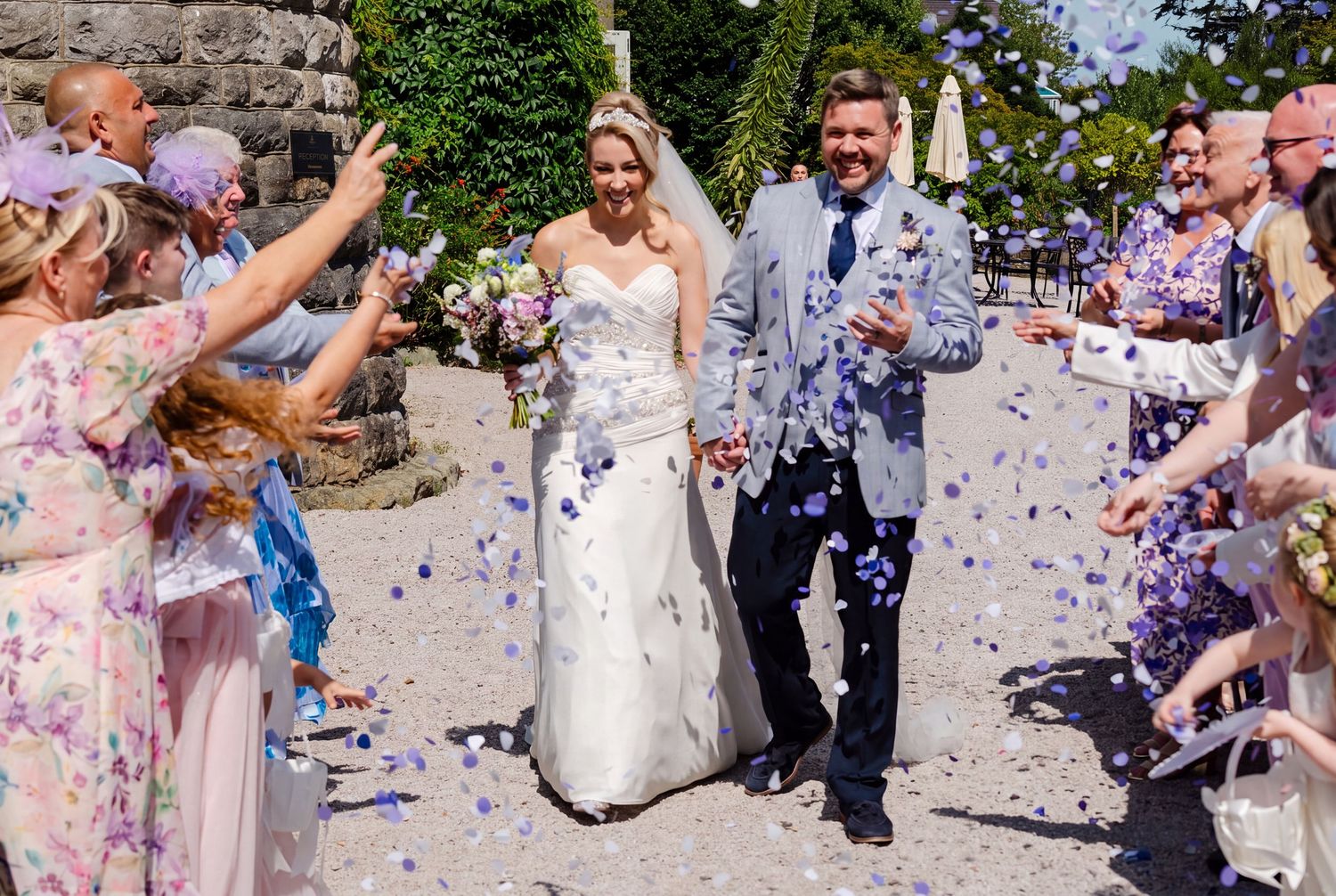 Bride and groom walk through confetti shower surrounded by cheering guests at outdoor wedding ceremony at Chateau Rhianfa, North Wales.
