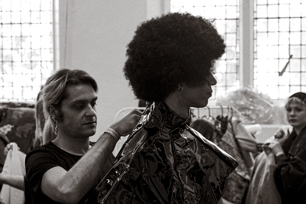 Fashion designer adjusting an ornate jacket on a model with an afro hairstyle in a studio setting.