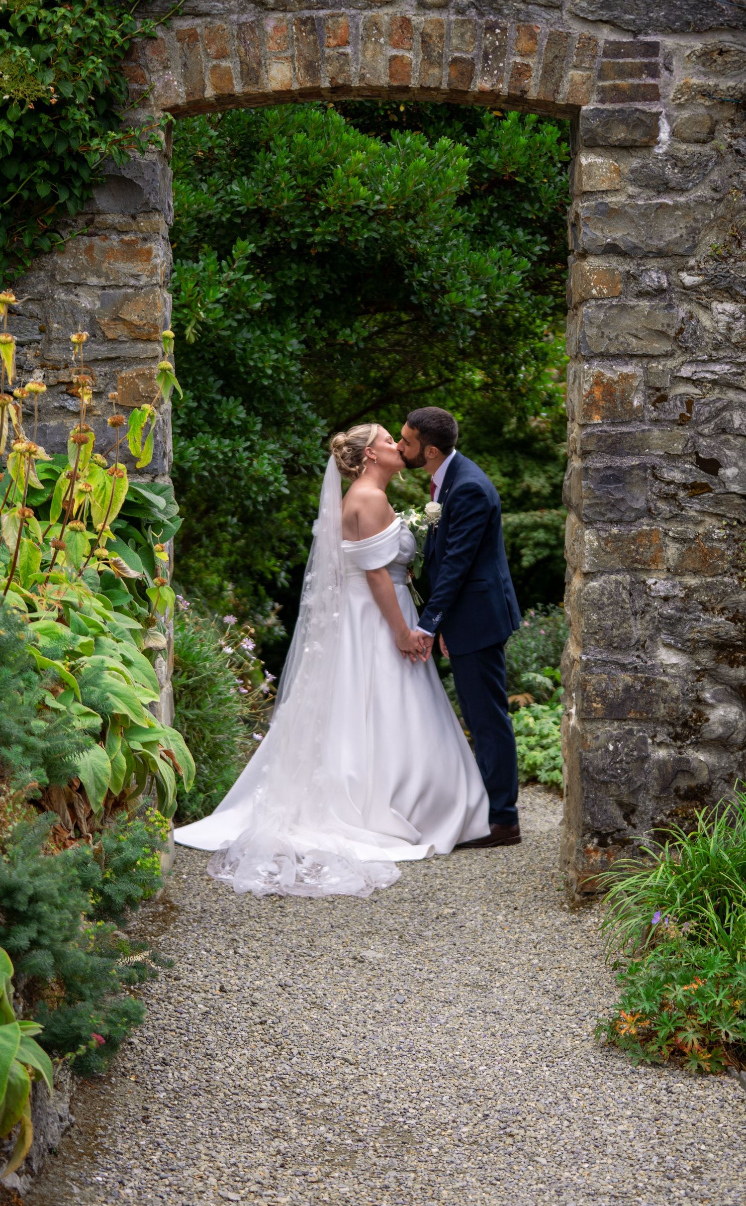 A bride and groom share a kiss under a stone archway surrounded by lush garden greenery on their wedding day.