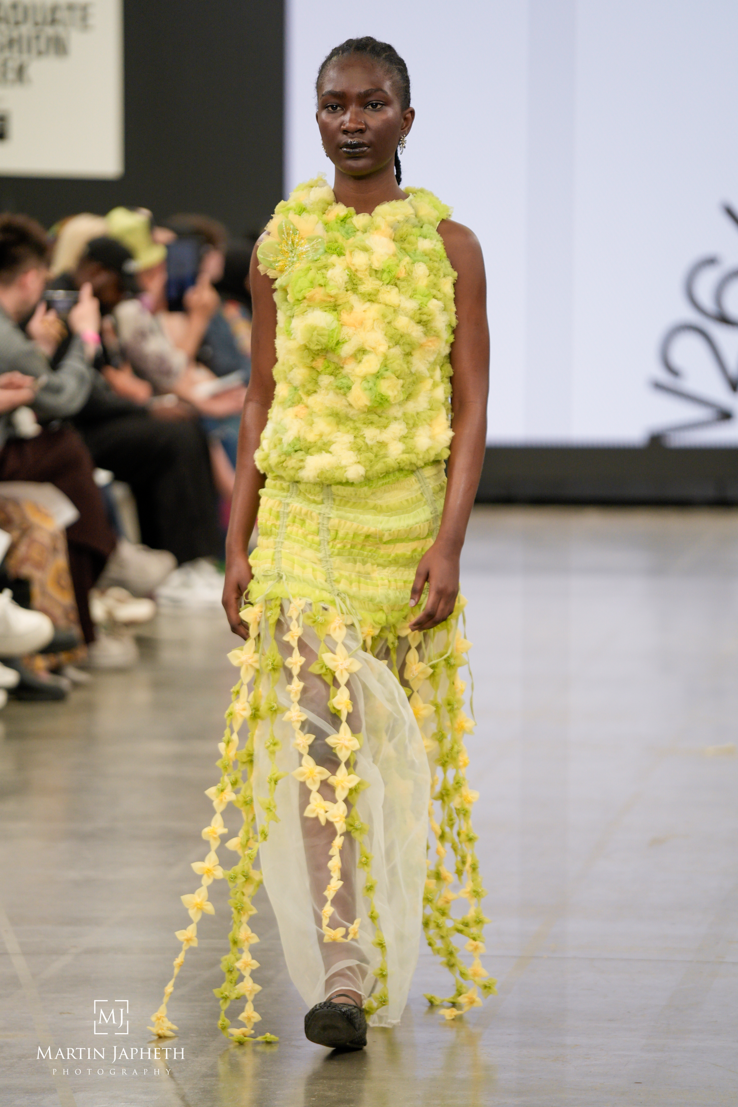 Model walks runway in yellow-green textured floral dress with sheer skirt at Graduate Fashion Week show. bMartin Japheth Photography