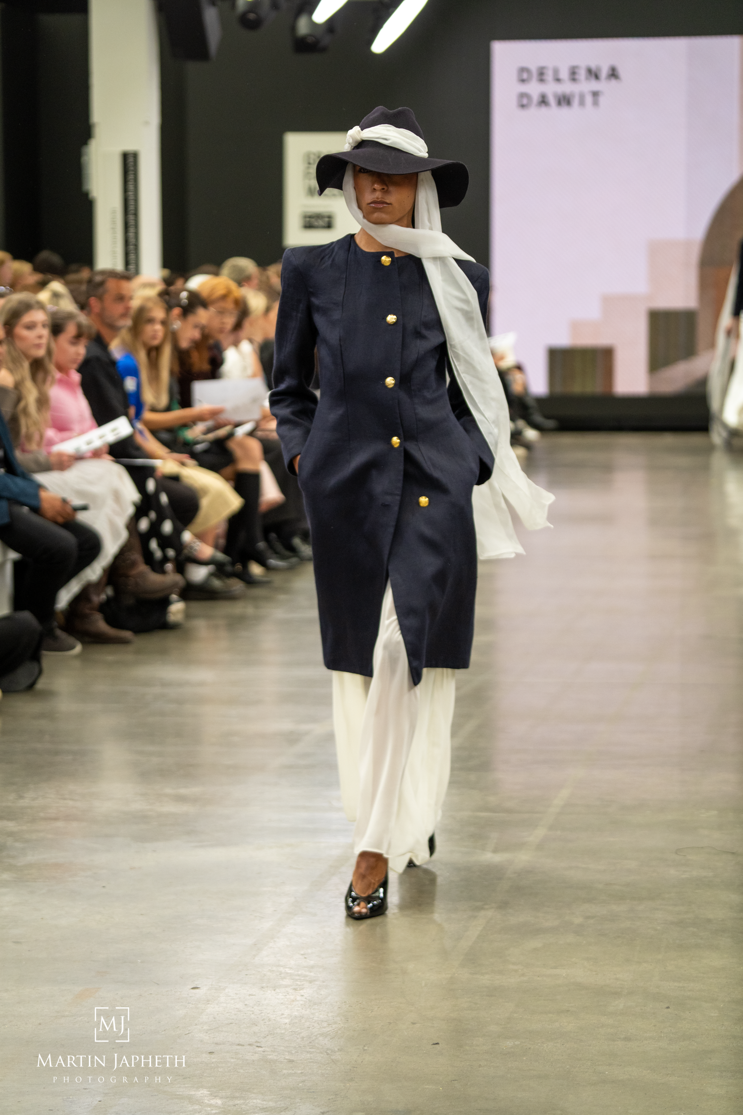 Model walks runway in navy coat with gold buttons, white wide-leg pants, and broad-brim hat at Delena Dawit fashion show. Martin Japheth Pho