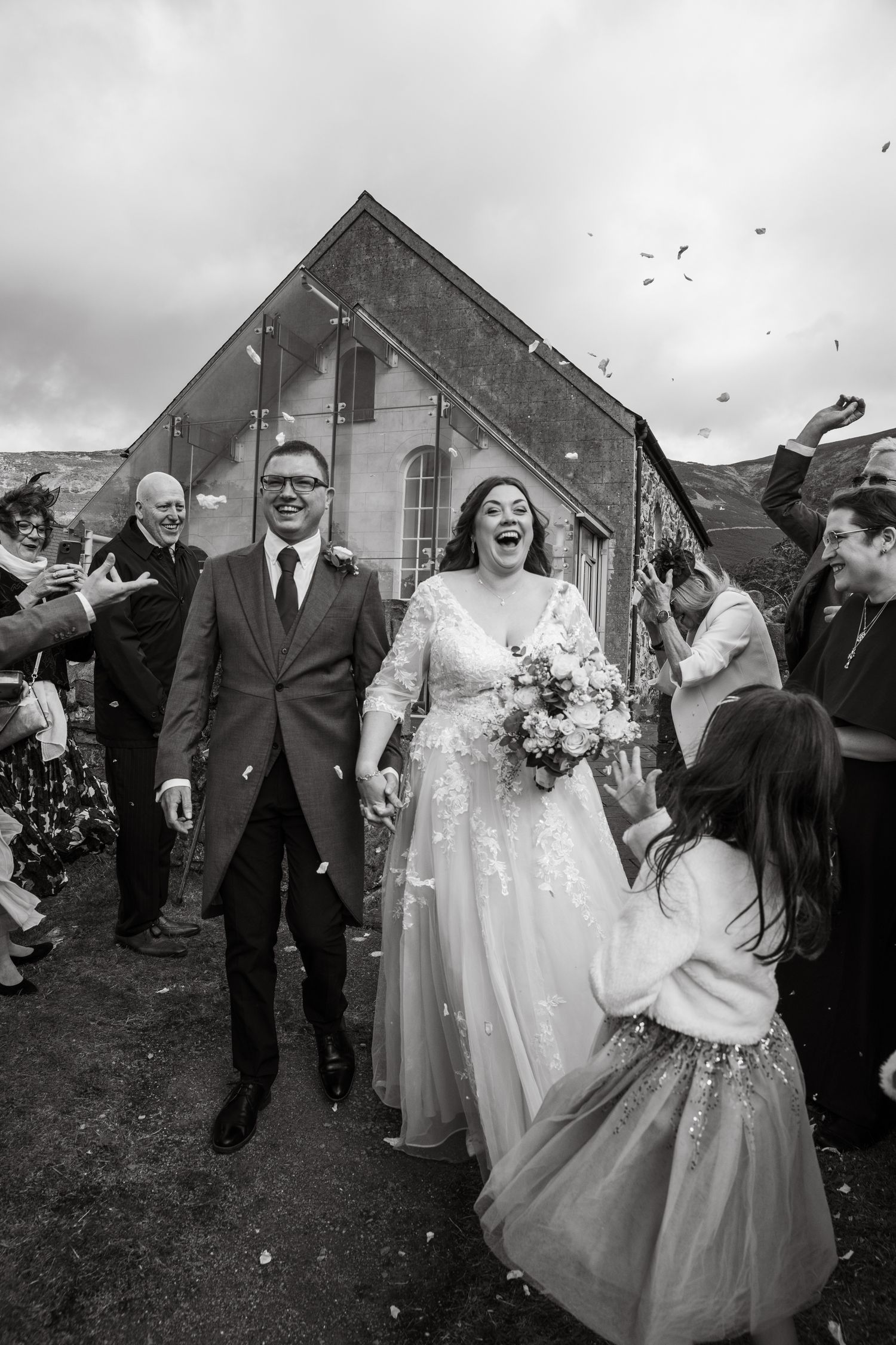 Black and white photo of a joyful bride and groom walking through confetti outside a church.
