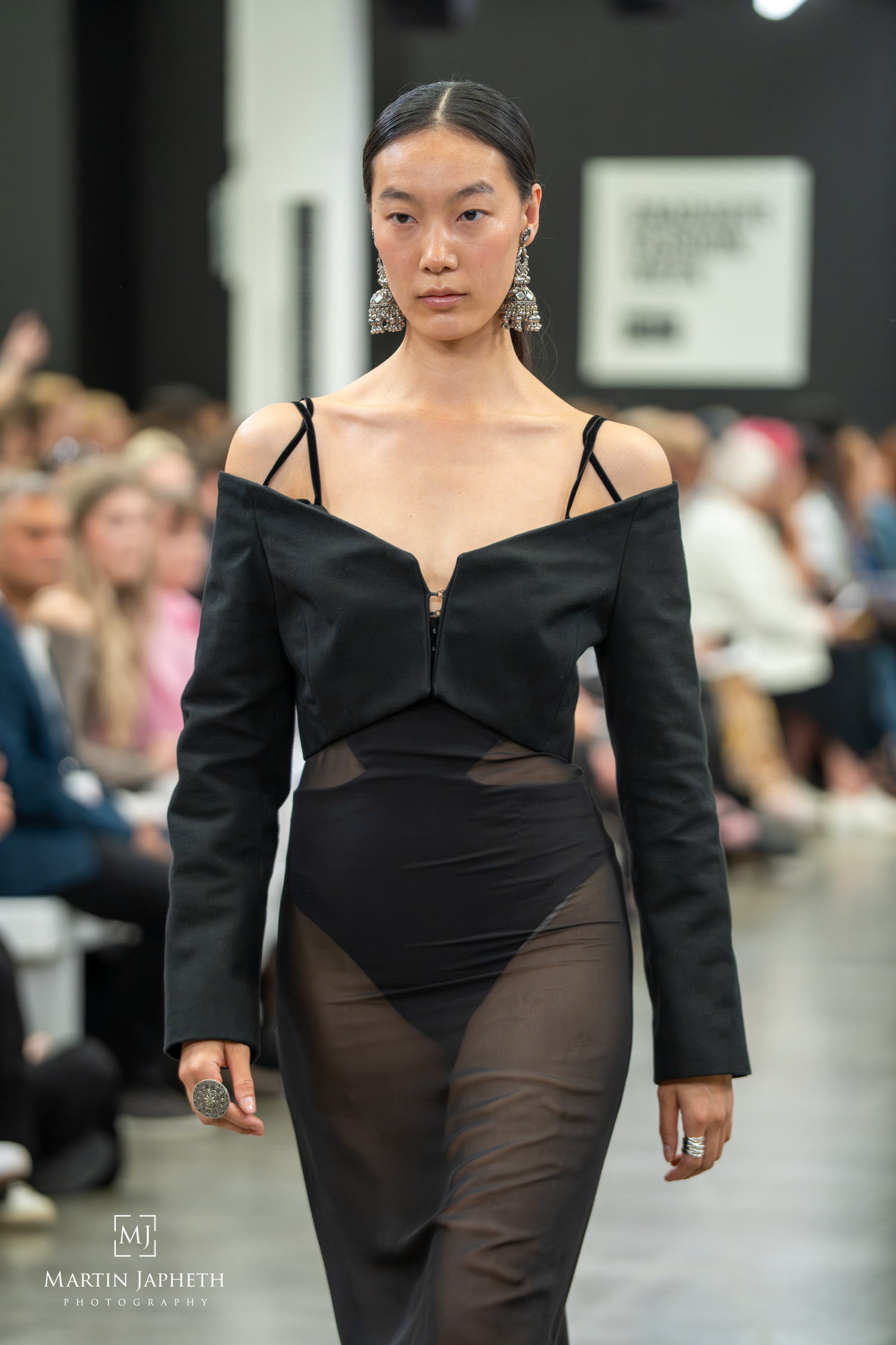 Model walks runway in black off-shoulder gown with sheer skirt and zip detail at Graduate Fashion Week 26. Martin Japheth Photography