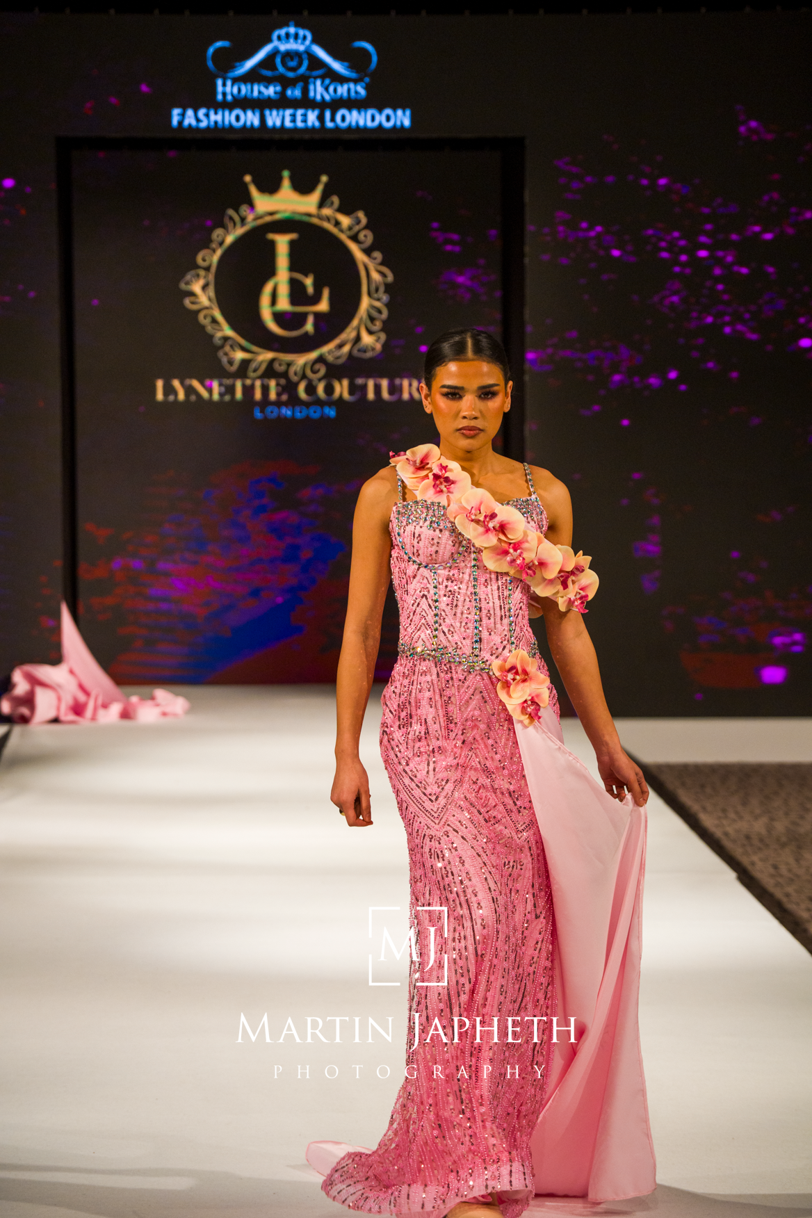 Model walks runway in pink floral one-shoulder evening gown during London Fashion Week show.