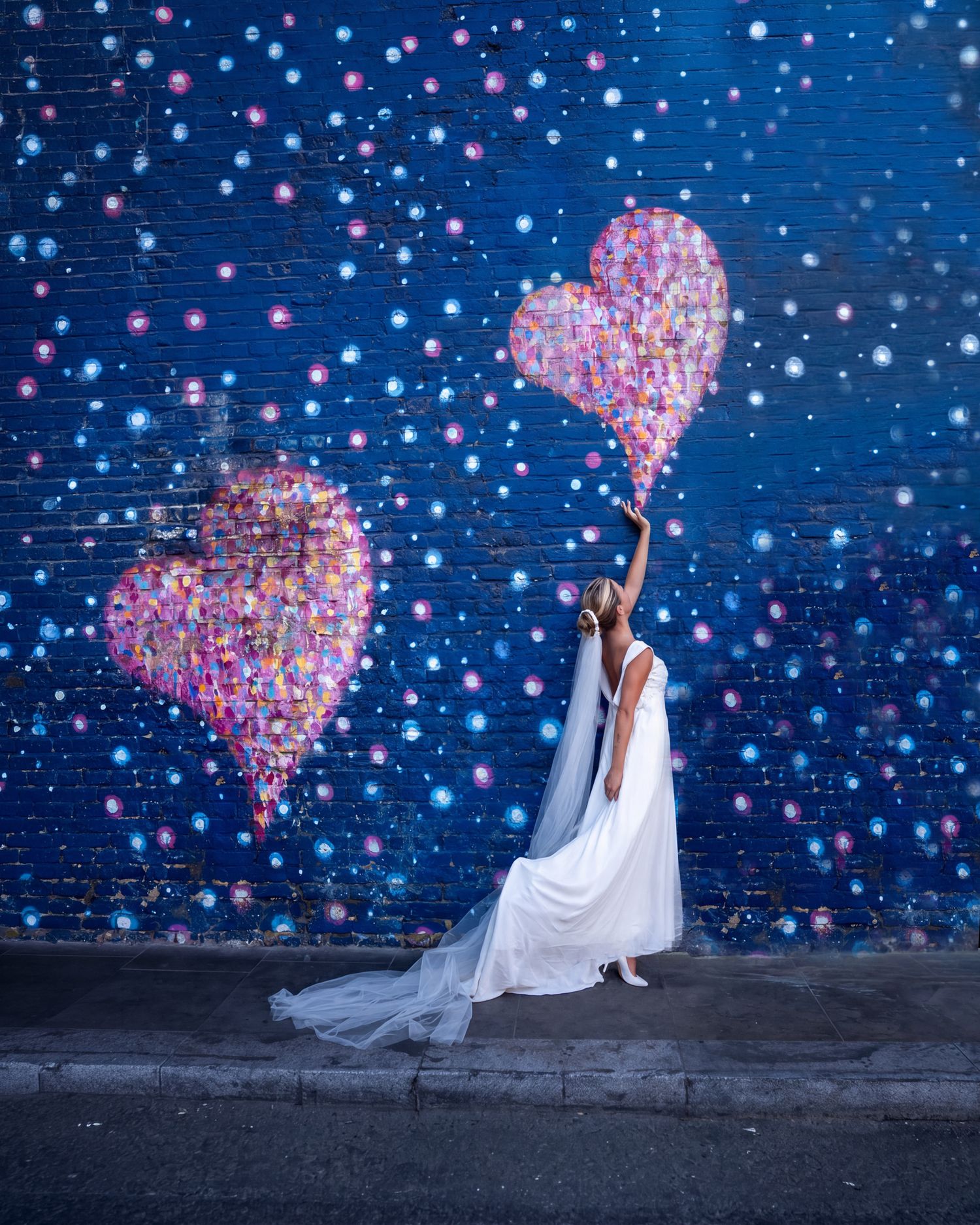 Bride in white gown and veil reaching up to touch a colorful heart mural on a blue brick wall at Borough Market, London with Martin Japheth.