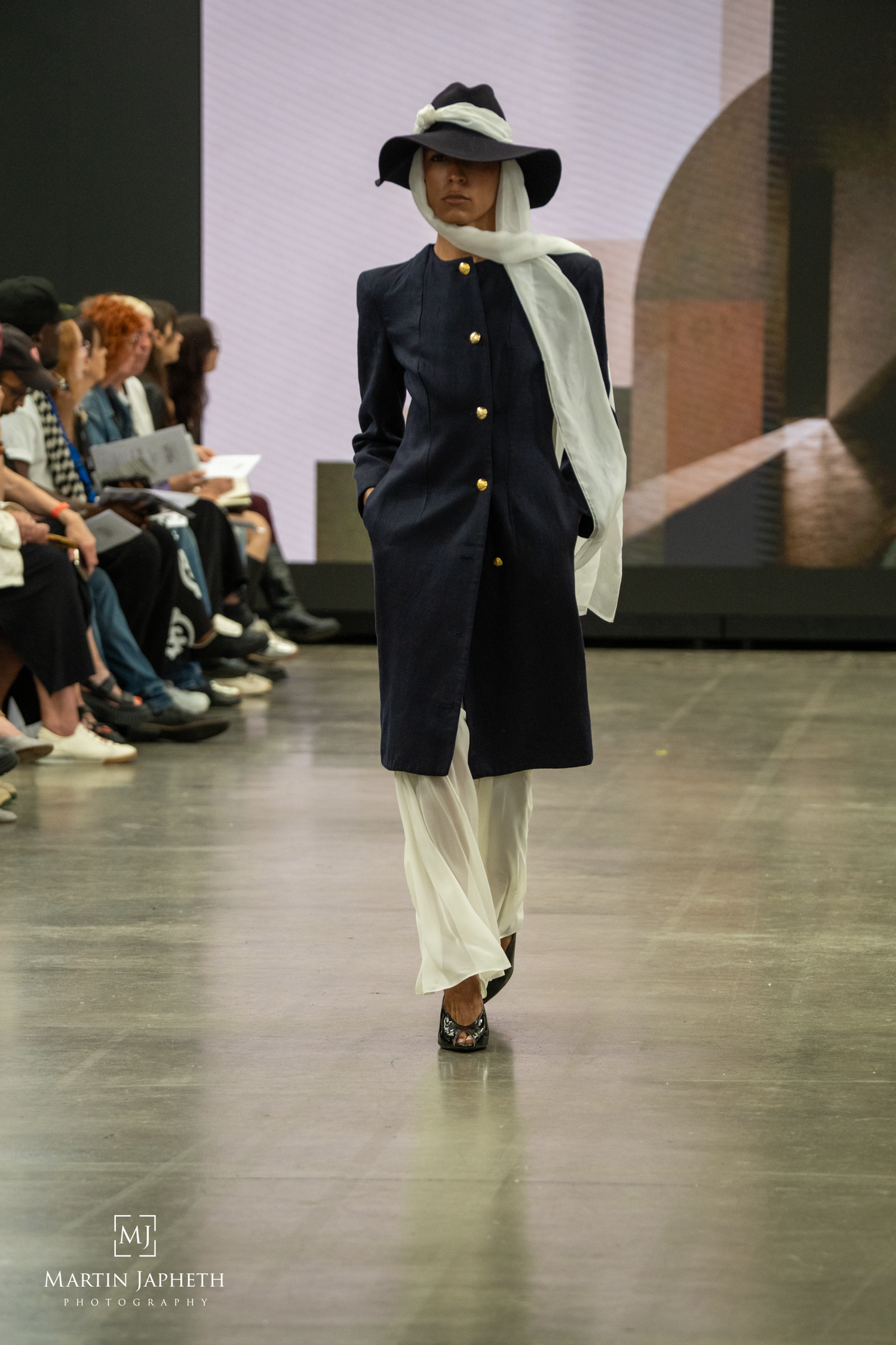 Model walks runway in navy coat with gold buttons, white wide-brim hat, and flowing white scarf at fashion show. Martin Japheth Photography