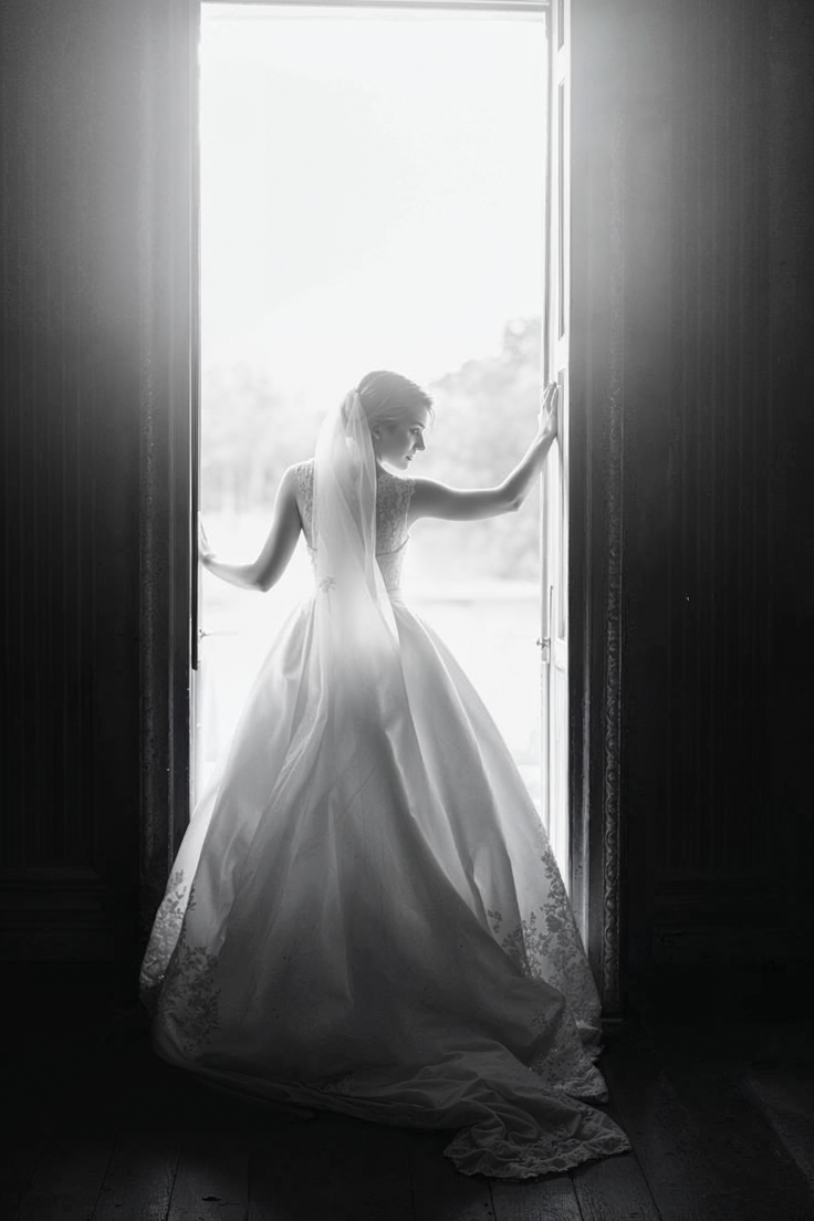 Bride in elegant ball gown and veil silhouetted in doorway with bright backlight, black and white wedding photo.