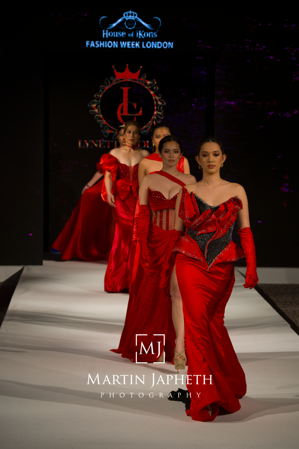 Models walk the runway in flowing red evening gowns during House of iKons Fashion Week London show.