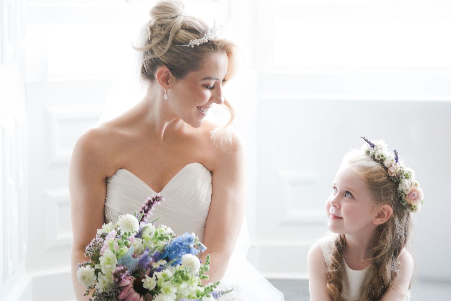 Bride in strapless gown holding floral bouquet smiles at flower girl wearing pink floral crown at wedding.