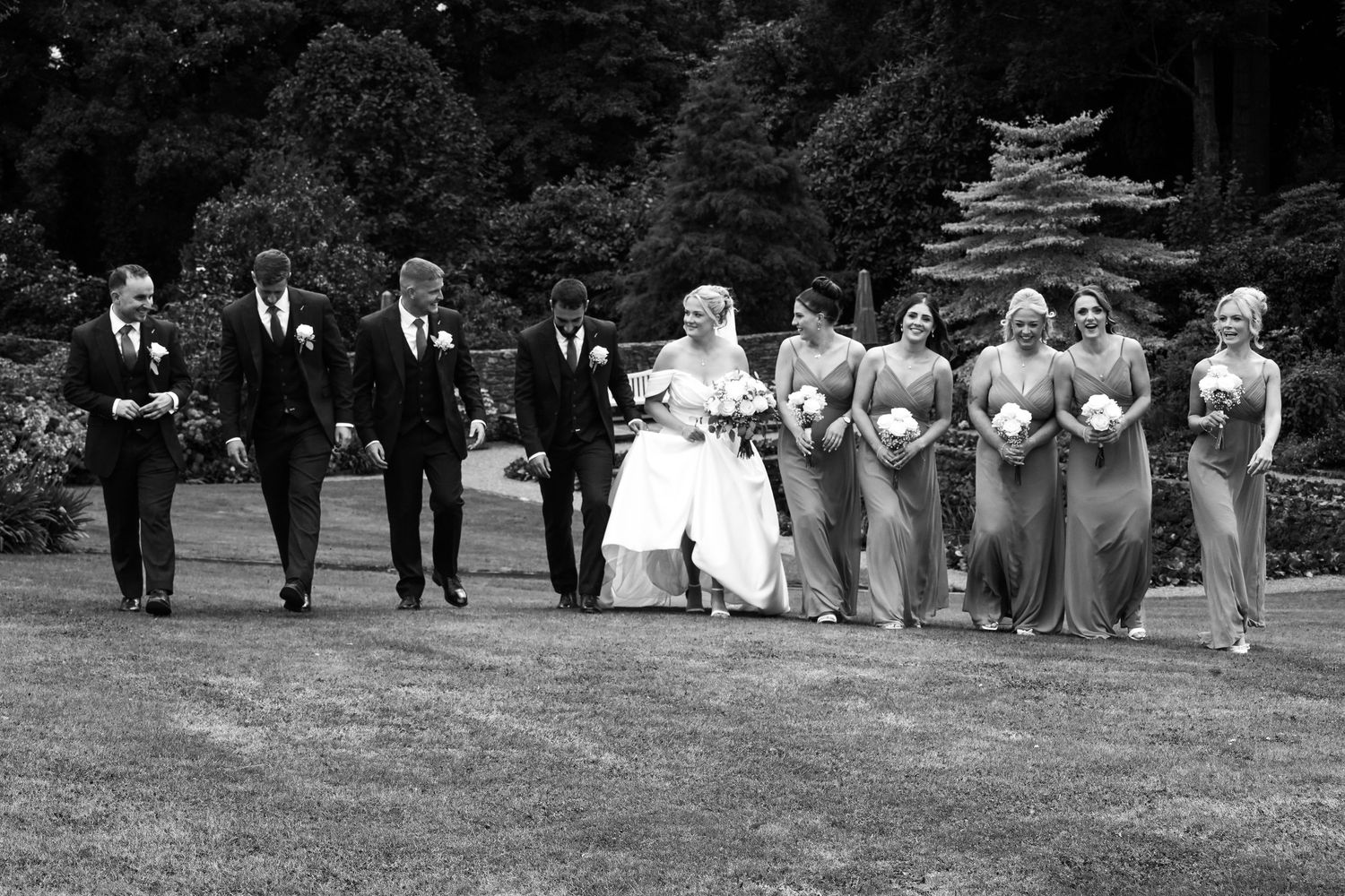 Black and white photo of a wedding party walking outdoors, bride in white gown with bridesmaids and groomsmen in formal attire.