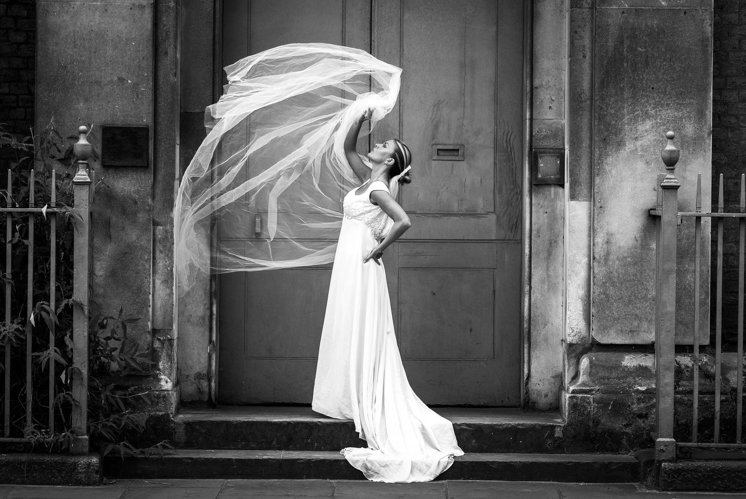 Black and white photo of a bride in a white gown with veil blowing in wind, standing before a vintage door.