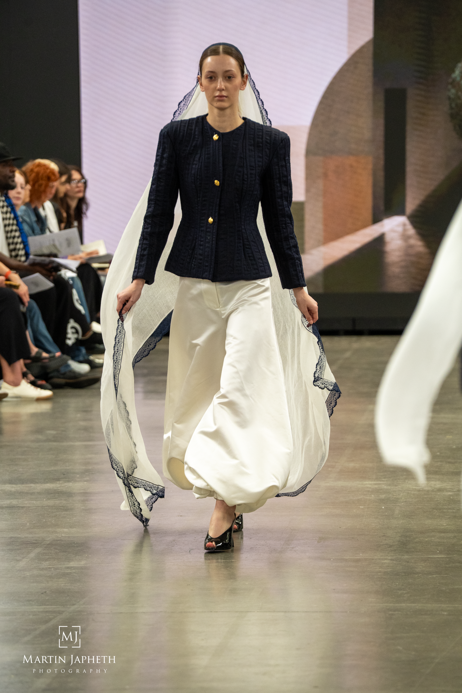 Model walks runway in navy structured jacket and flowing white skirt with lace-trimmed veil at fashion show. Martin Japheth Photography