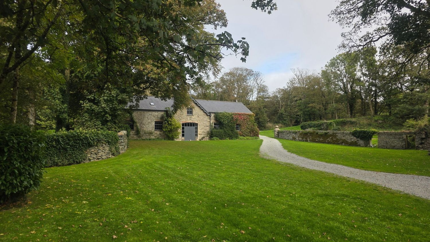 Stone cottage with slate roof surrounded by mature trees and a manicured green lawn with a winding gravel driveway.