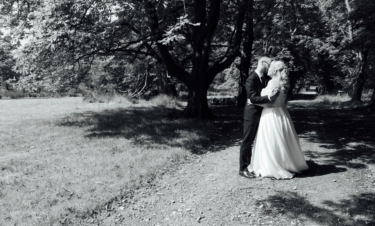 Black and white photo of a bride and groom embracing under a large tree in a park on their wedding day.
