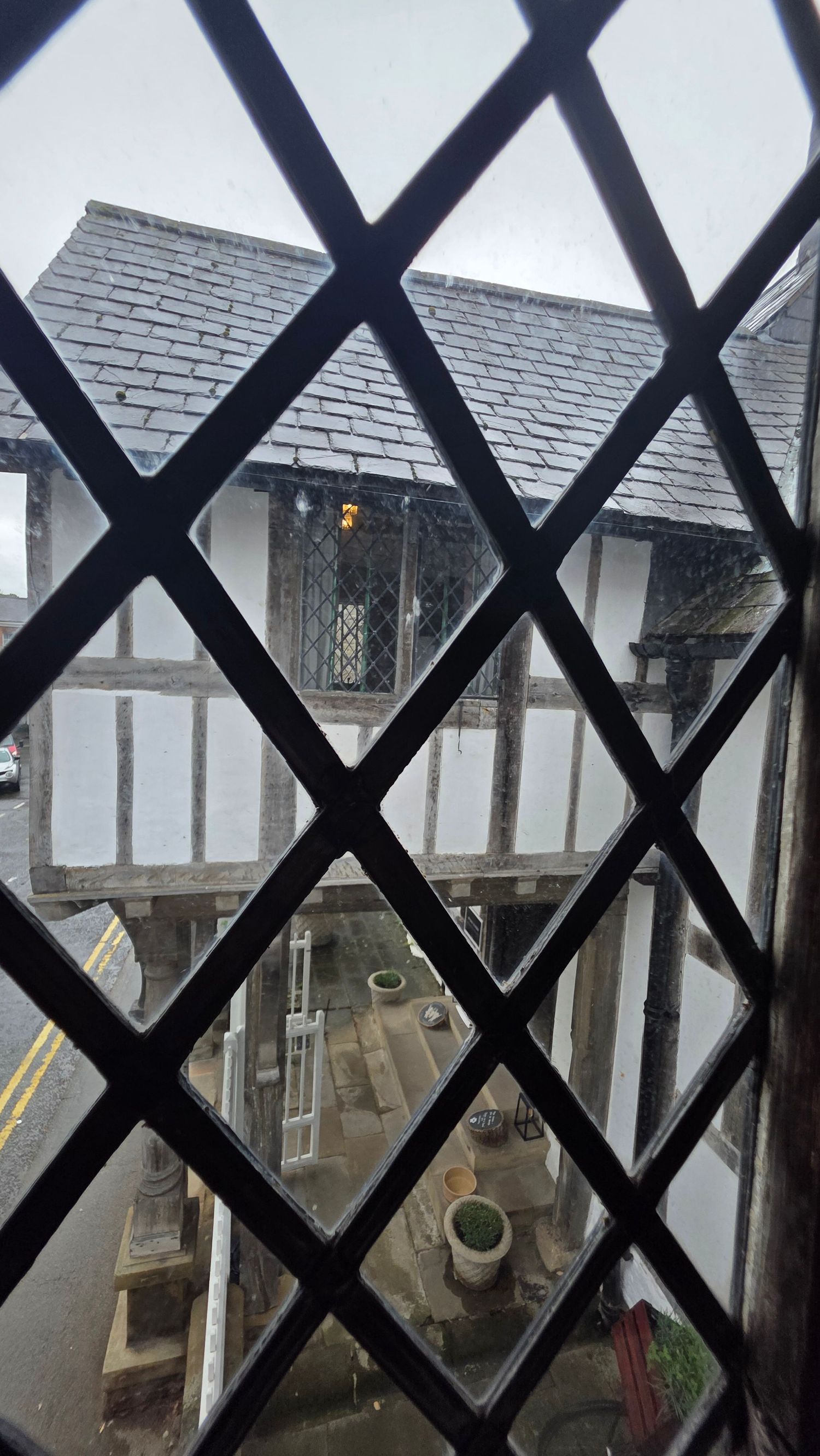 View through diamond-patterned leaded glass window of a historic Tudor timber-framed building on a rainy day.