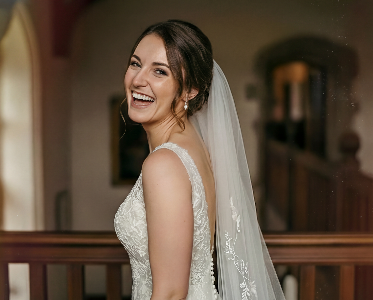 A bride in a white lace wedding dress laughs joyfully while wearing a flowing veil during her wedding day.