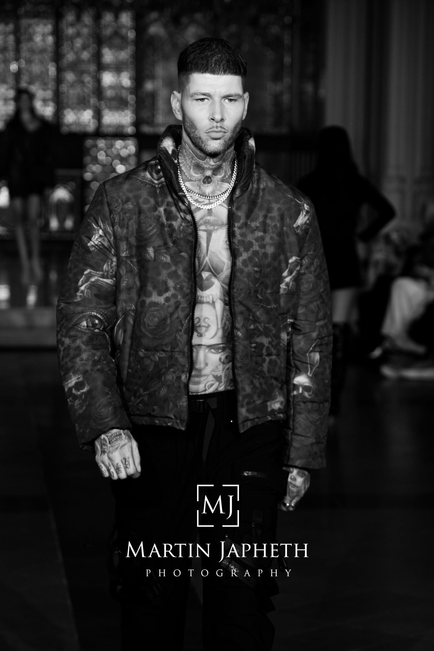 Black and white fashion photograph of a model wearing a distressed leather jacket and showing visible tattoos on the runway.