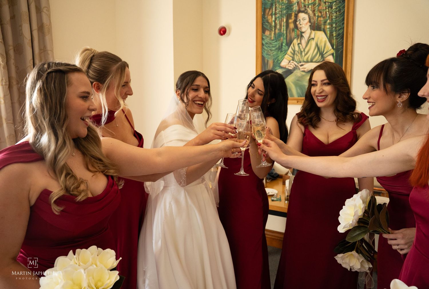 Bride in white gown toasting champagne with bridesmaids in red dresses during wedding preparations. Martin Japheth Photography