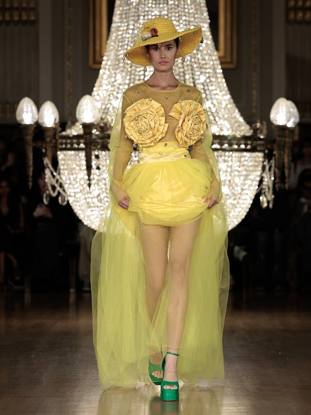 Model walks runway in yellow ruffled dress with sheer lime green overlay at fashion show under crystal chandeliers.