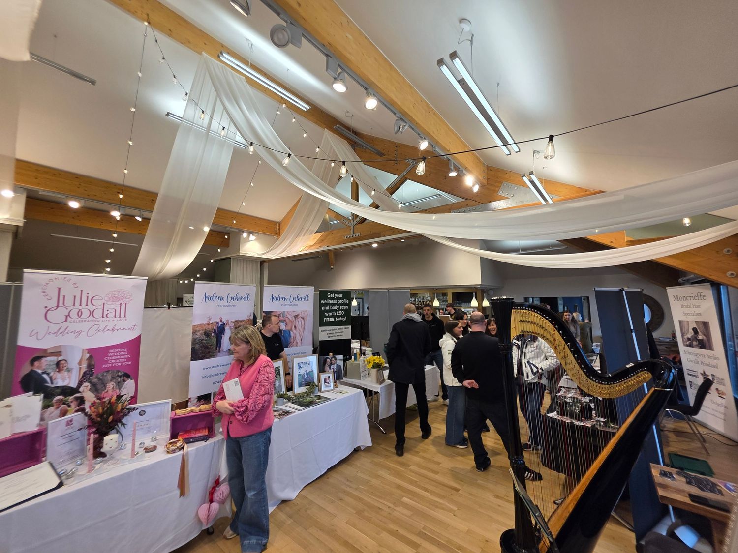 A panoramic view of an indoor event space with display tables, banners, and attendees browsing exhibits at a Nant Gwrtheyrn Wedding Fayre.