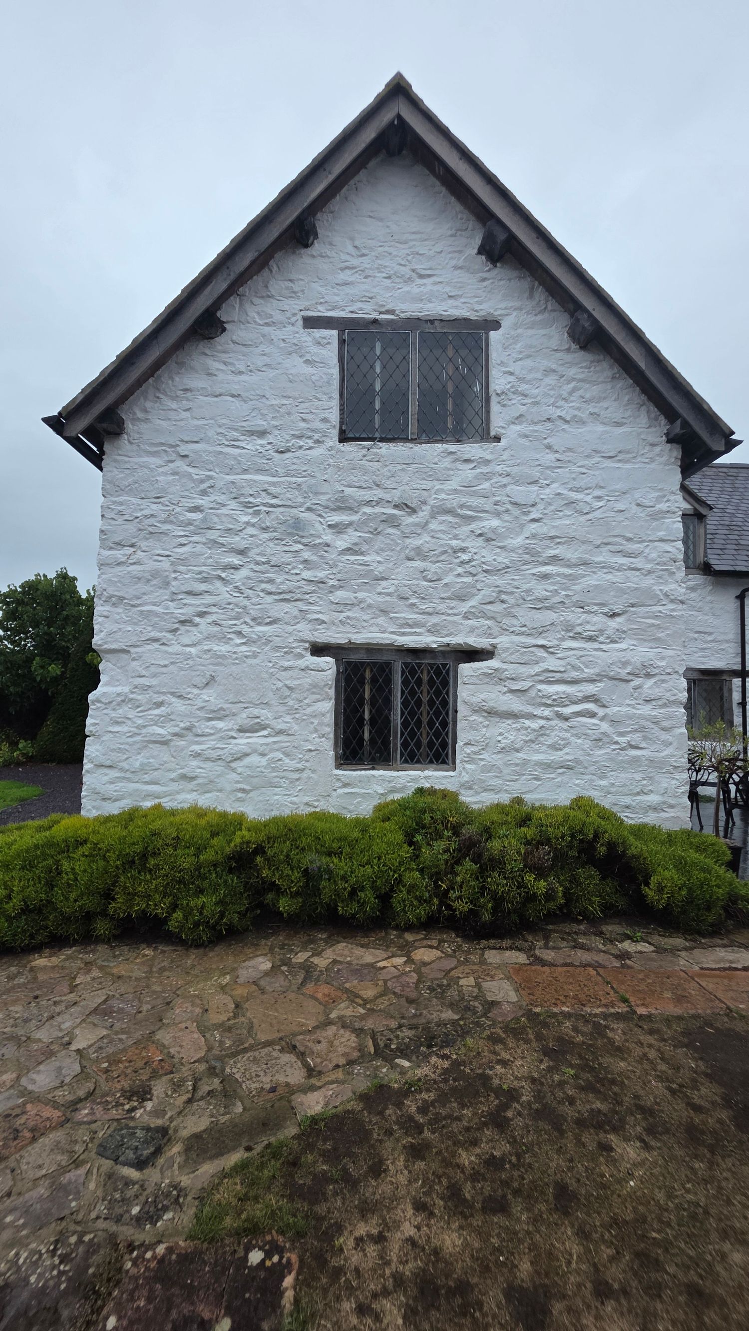 White-painted stone cottage with leaded windows, gabled roof, and neatly trimmed shrubs on a cobblestone path.