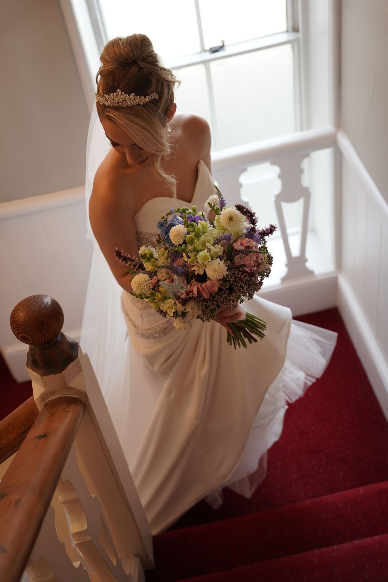 Bride in white strapless gown holding colorful bouquet descends red-carpeted staircase, hair up with floral tiara.wedding at Chateau Rhianfa