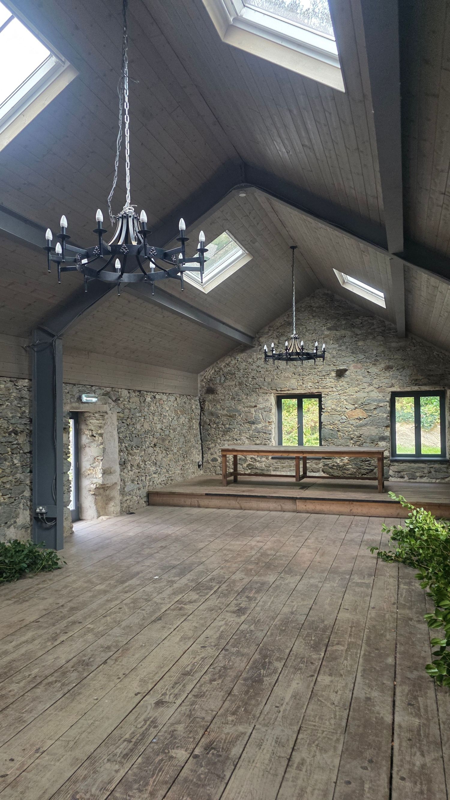 Rustic stone barn interior with vaulted wood ceiling, skylights, black iron chandeliers, wide plank floor and a raised platform with a long 