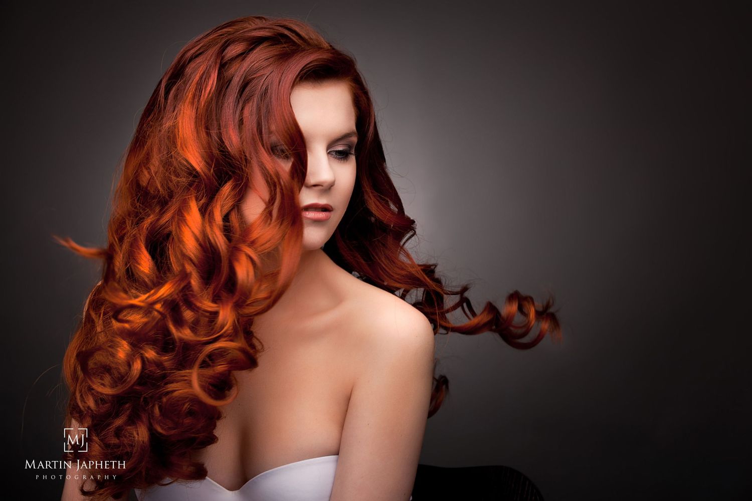 Studio portrait of a woman with voluminous red curly hair wearing a white strapless top against a dark background.