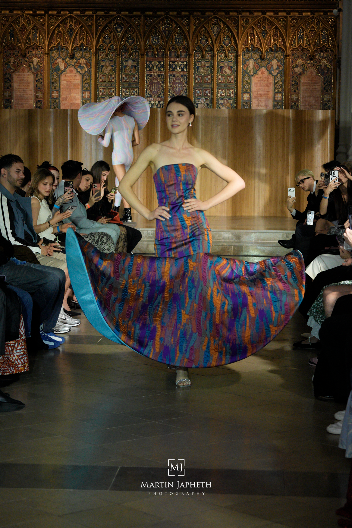 Model walks runway in colorful strapless ikat gown at fashion show, audience seated alongside catwalk.