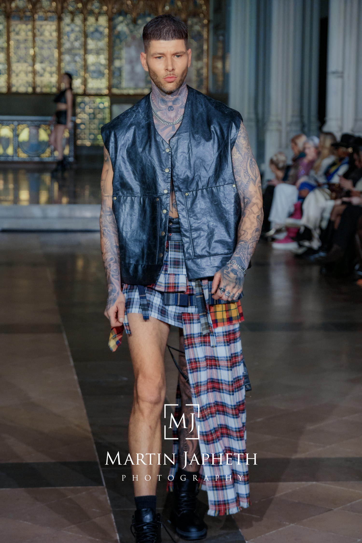 Fashion model wearing black leather vest and plaid kilt walks runway during fashion show presentation.