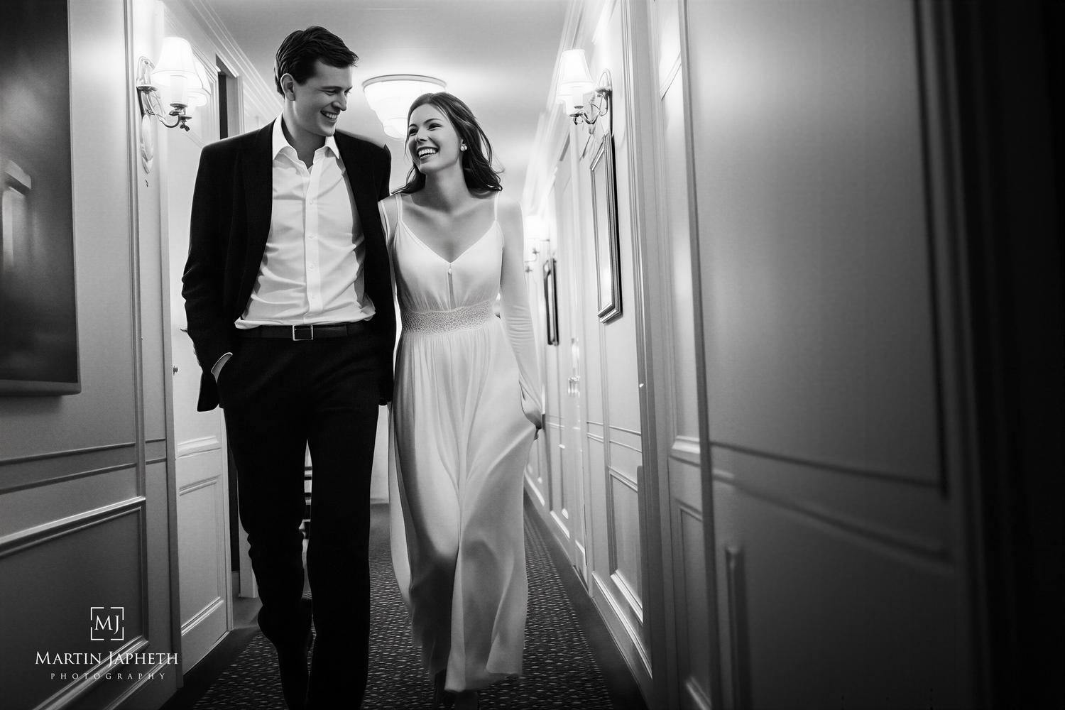 Black and white photo of a smiling couple in wedding attire walking together down an elegant hotel hallway.