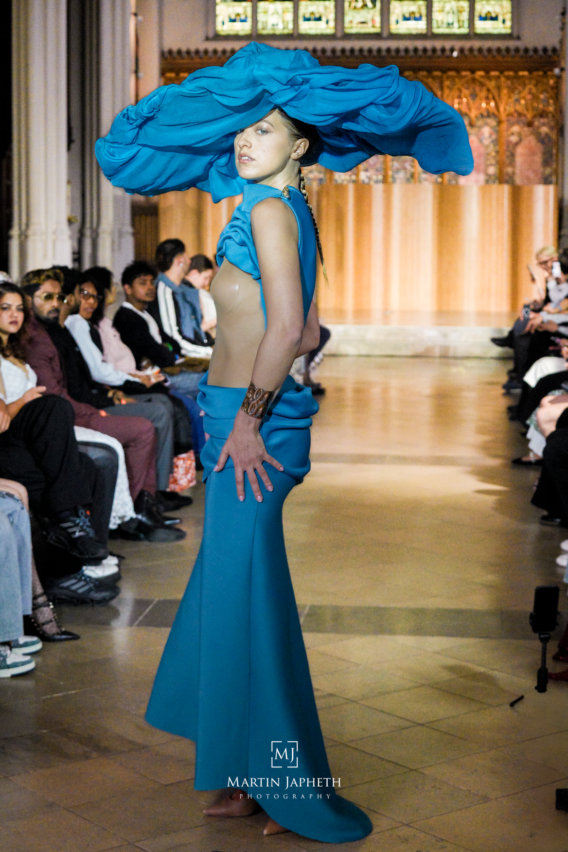 Model walks runway in teal backless gown with dramatic oversized hat at fashion show.