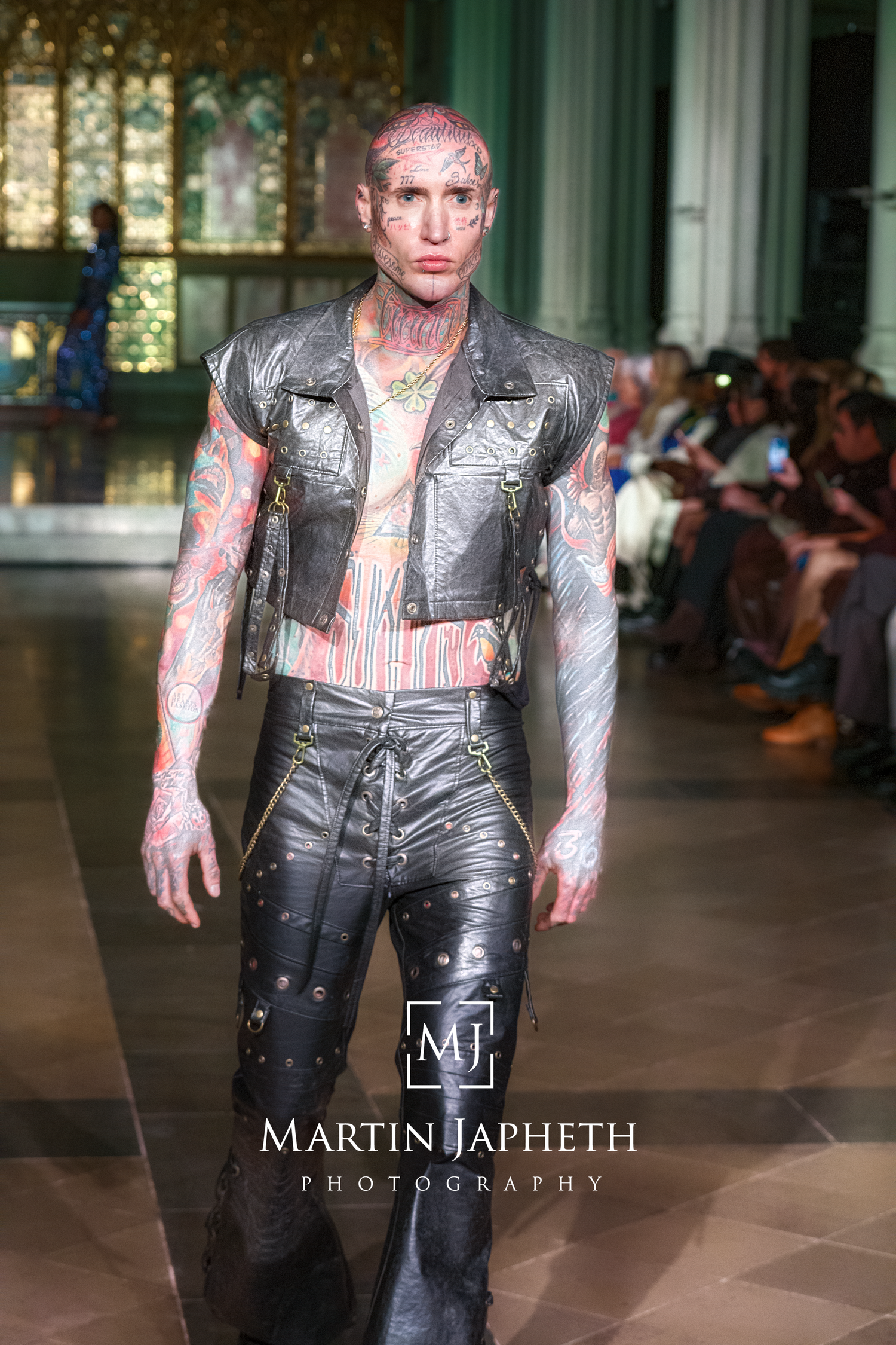 Model walks runway in edgy black leather vest and pants with tattoo-style body art at fashion show.