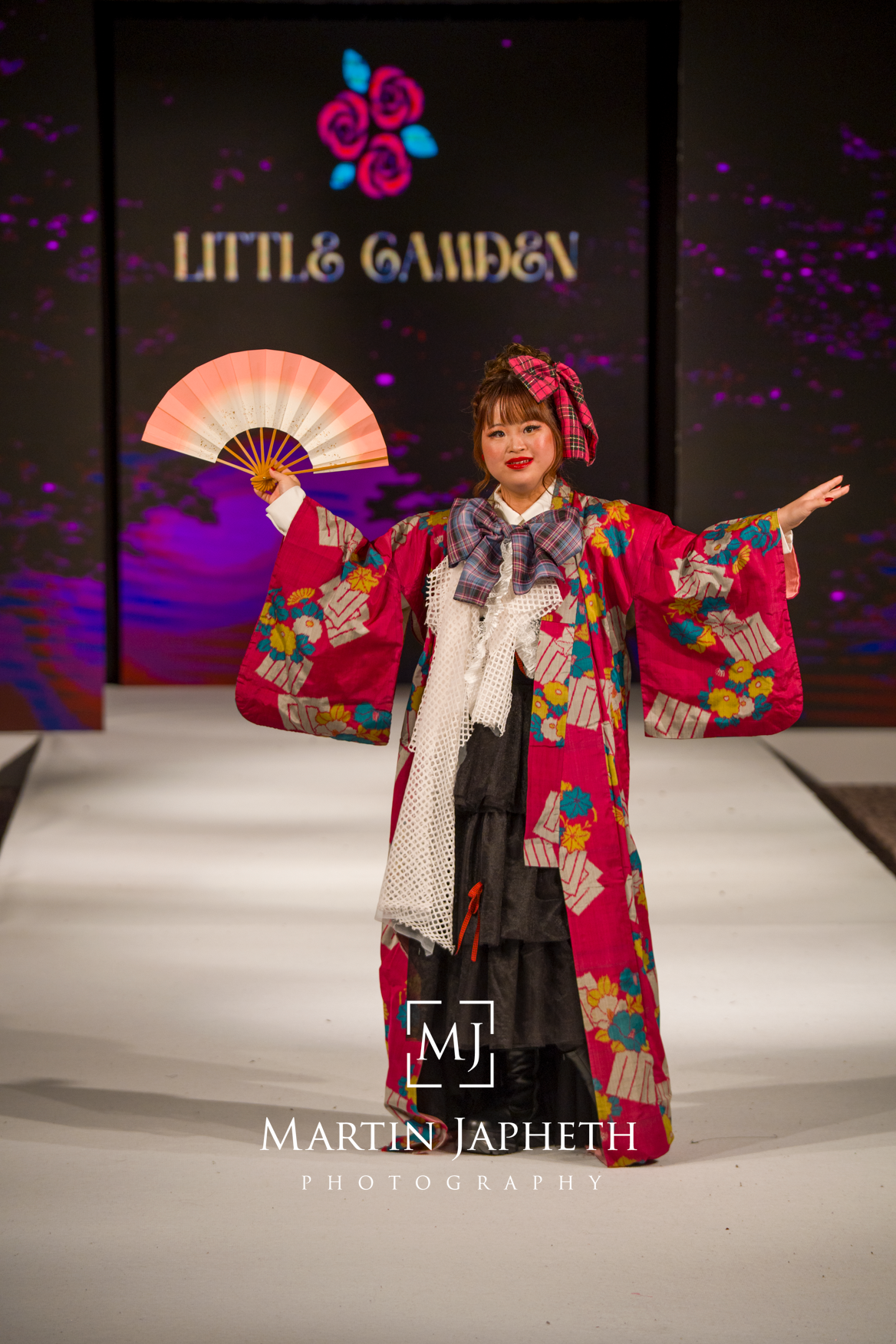 Fashion model wearing colorful kimono-inspired outfit with decorative fans performs on illuminated runway stage.