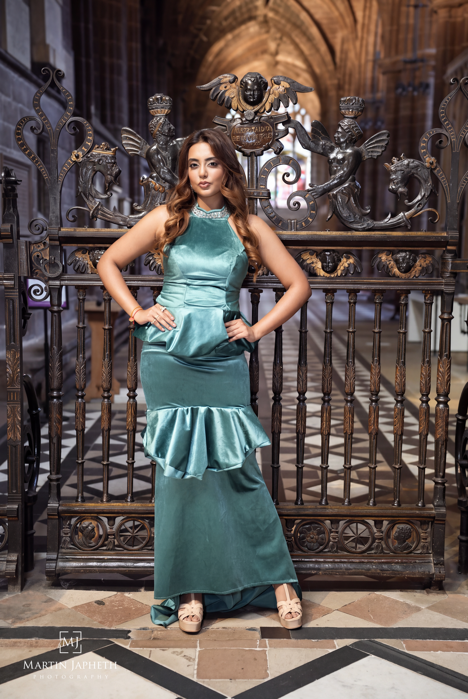 Woman in teal satin mermaid gown poses before ornate iron gates in a grand cathedral setting.