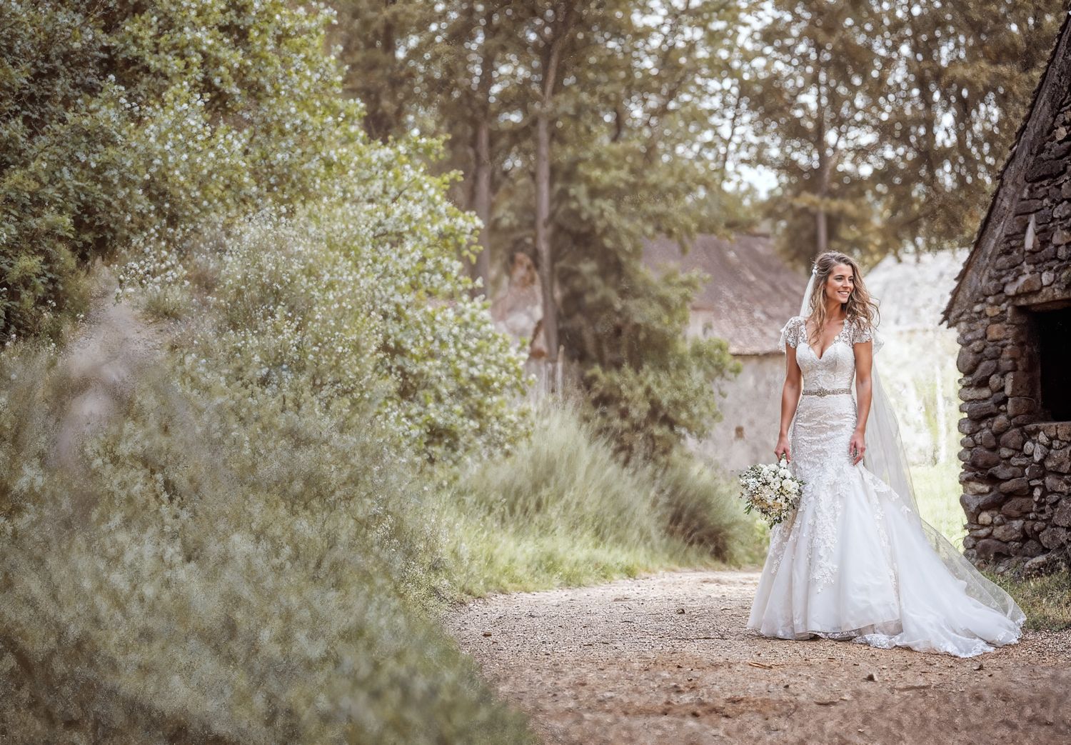 Bride in lace mermaid wedding dress holding bouquet smiling outdoors near stone building surrounded by lush greenery.