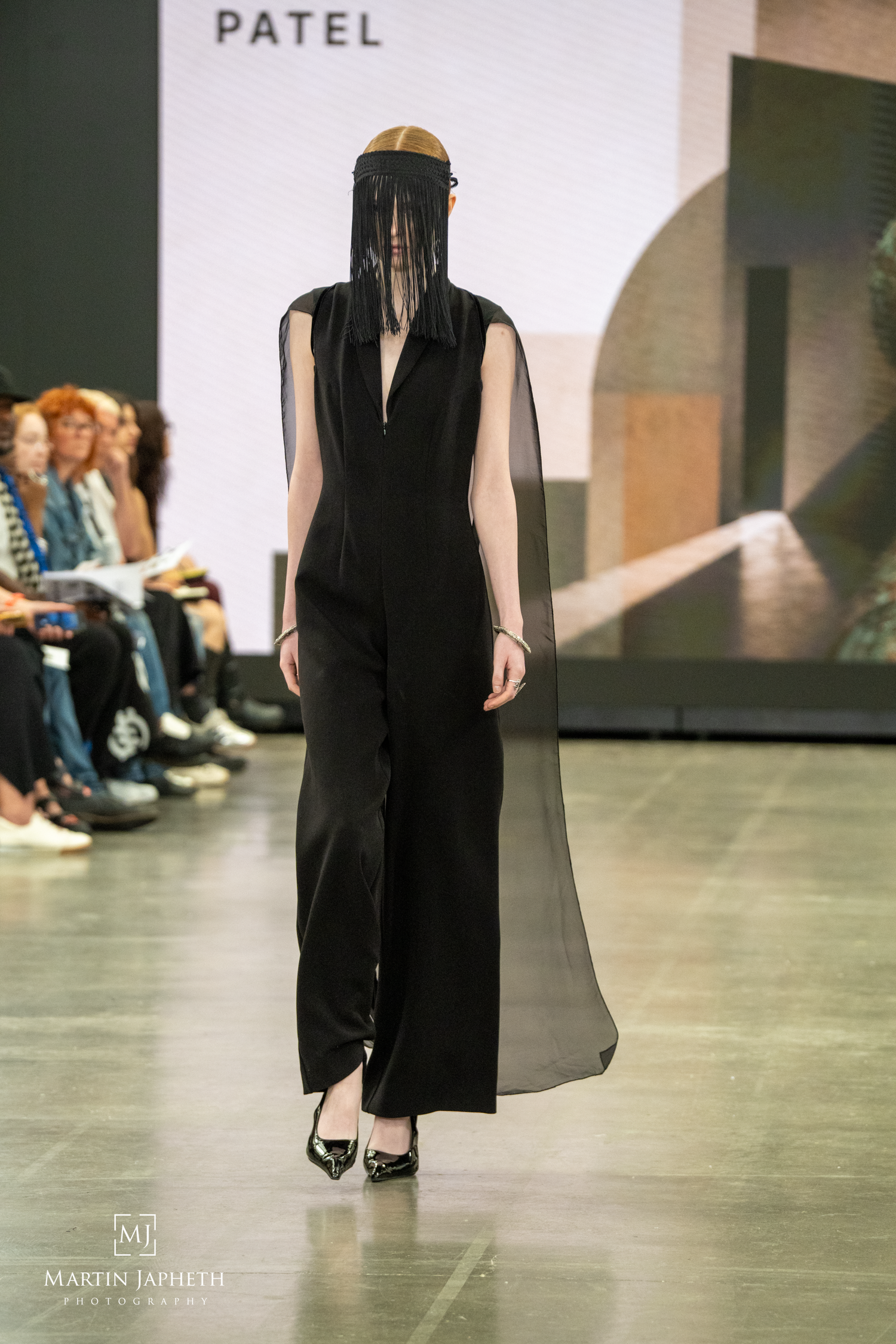 Model walks runway in black sleeveless jumpsuit with sheer cape and veiled headpiece at Patel fashion show. Martin Japheth Photography