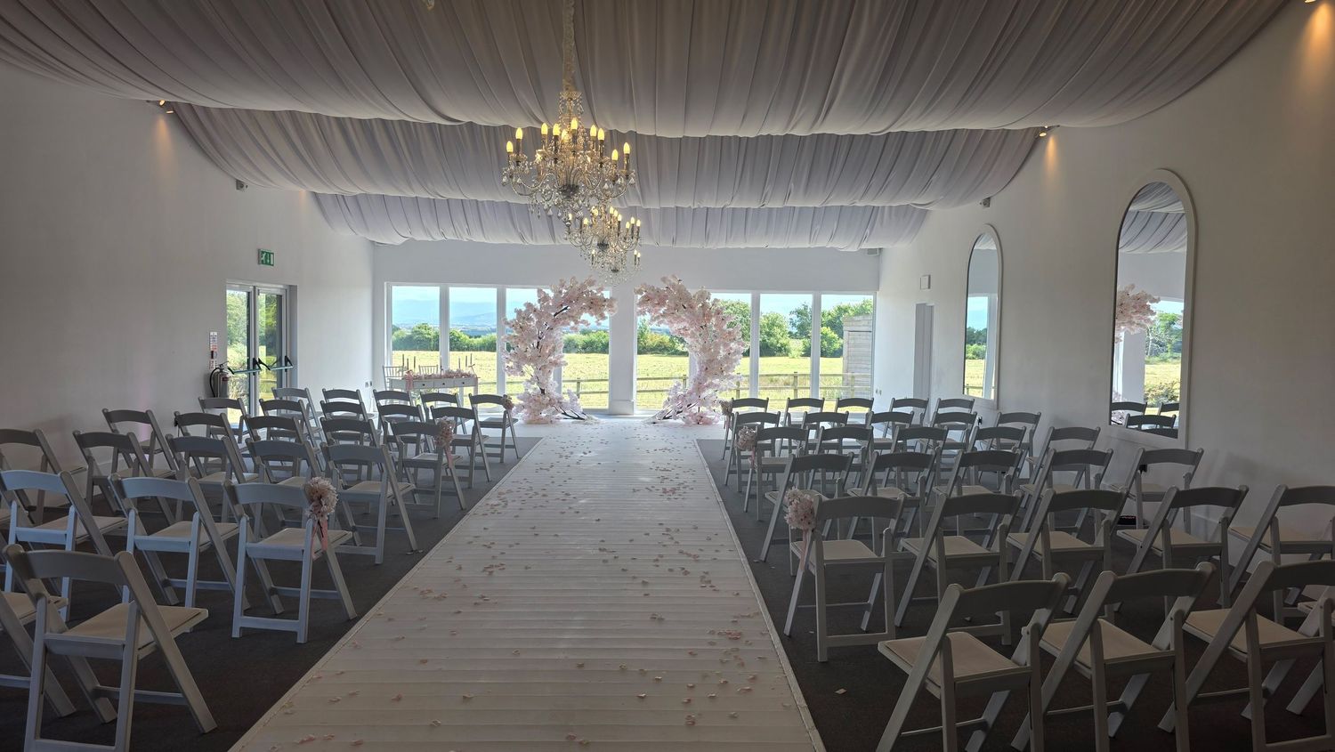 Elegant wedding ceremony venue with white draped ceiling, chandelier, floral arch, and rows of white folding chairs on a petal-lined aisle.