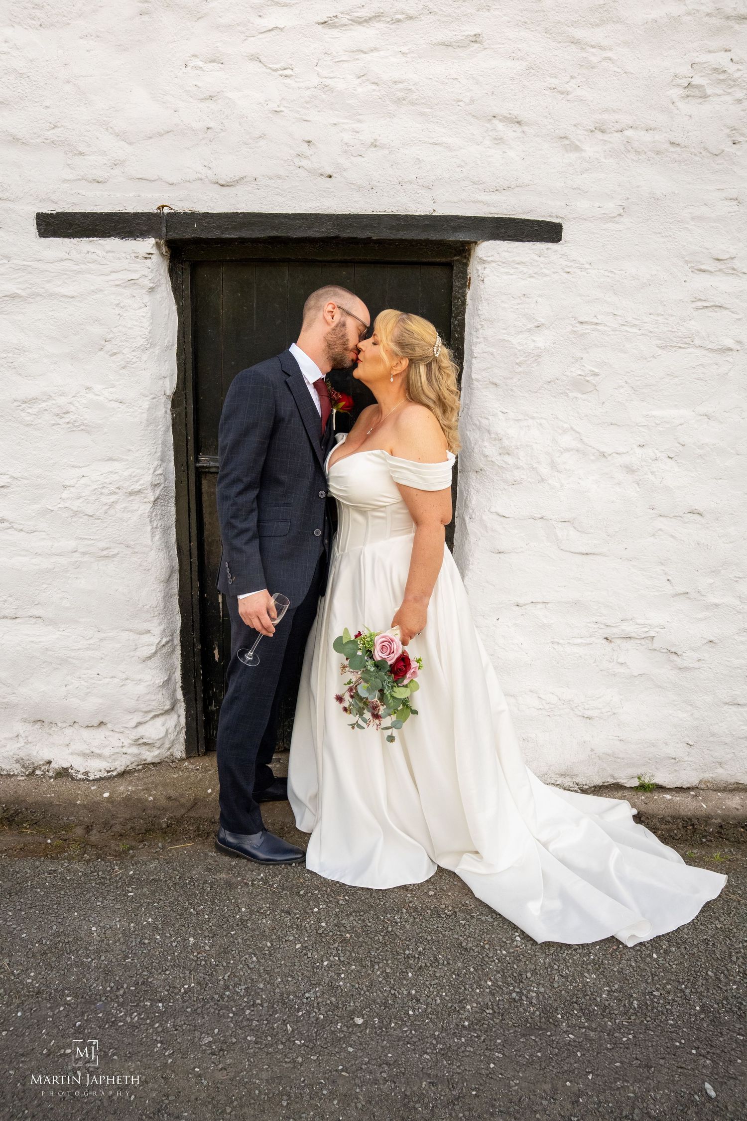 Bride and groom share a kiss in front of a rustic white stone wall with a dark wooden door on their wedding day.