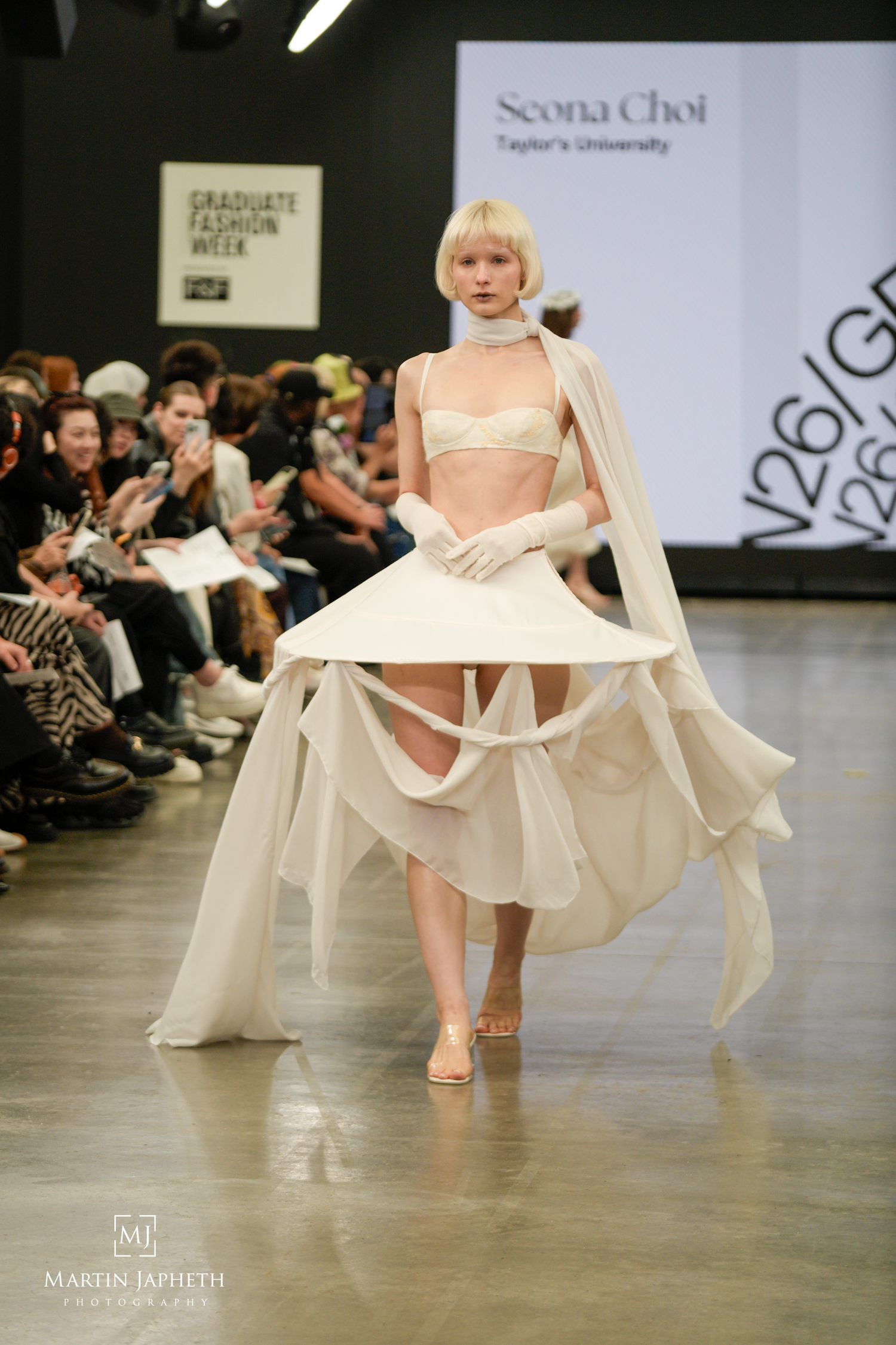 Model walks runway at Graduate Fashion Week in Seona Choi Taylor's University white avant-garde ensemble with cape. Martin Japheth