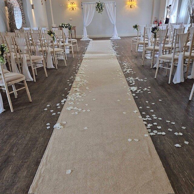 Elegant wedding ceremony aisle with ivory petal runner, gold chiavari chairs, and floral arch backdrop in a bright venue.