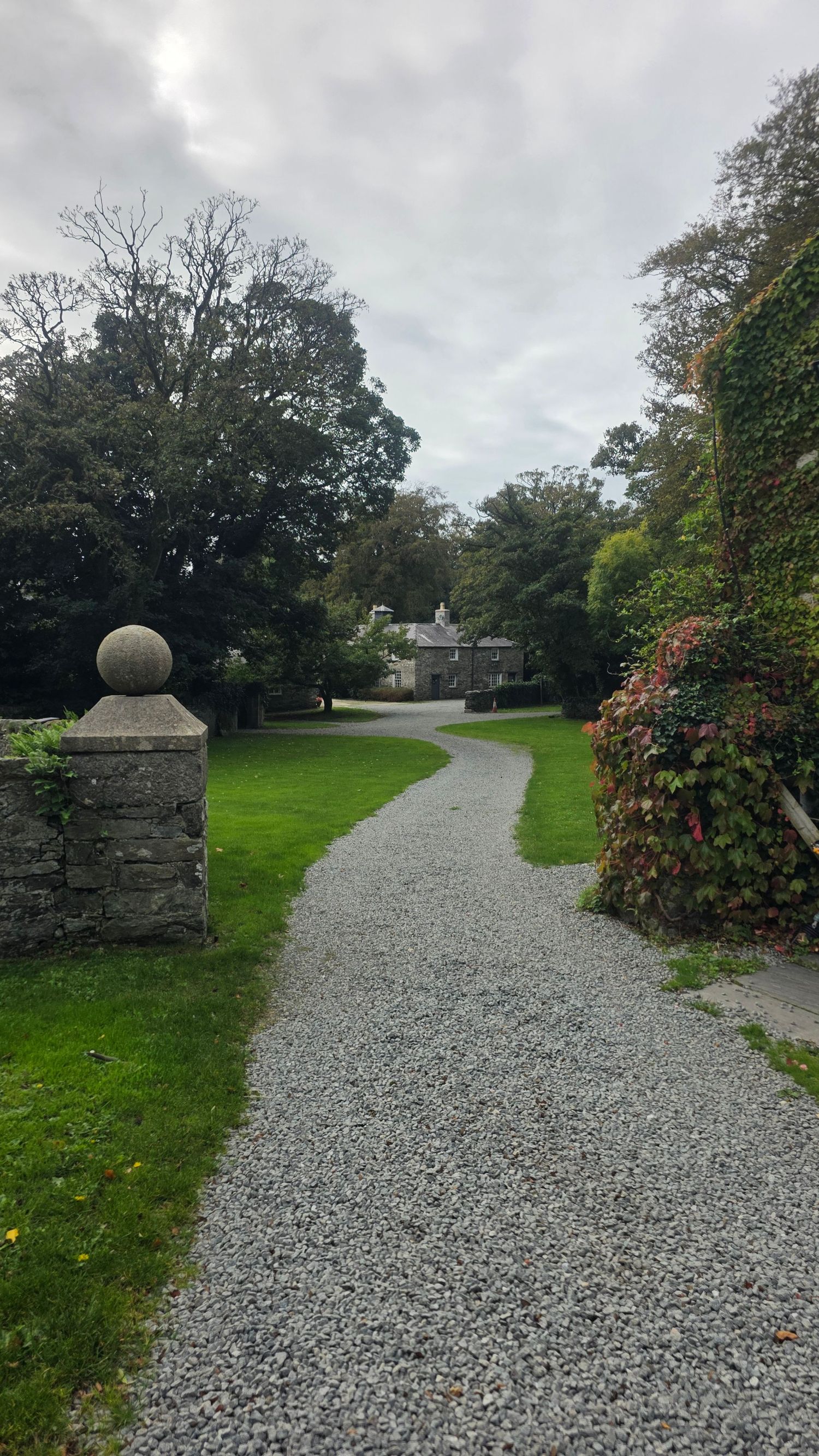 Curving gravel path leads past a stone pillar with a ball finial and ivy-covered wall toward a distant manor house surrounded by trees.