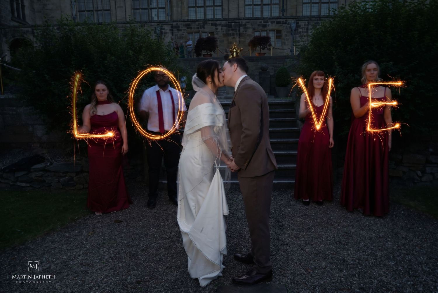 Wedding couple kissing while bridesmaids in red dresses hold sparklers spelling LOVE at dusk outdoor venue.