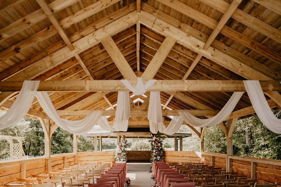 Rustic open-air wedding chapel with wooden beams, white fabric draping, and floral arch altar surrounded by wooden pews.