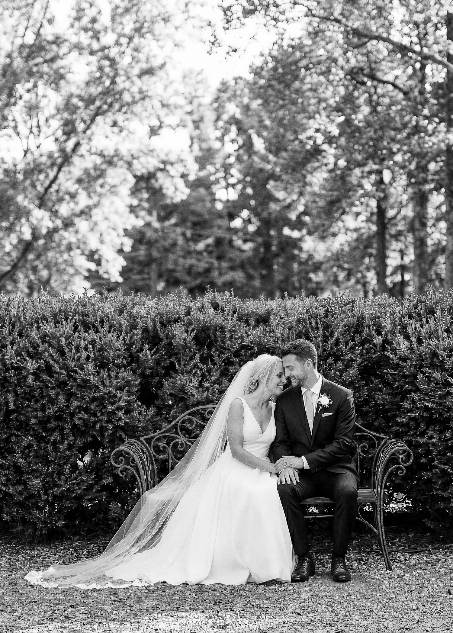 Wedding couple seated on ornate garden bench surrounded by lush greenery. Wedding Photographer Martin Japheth