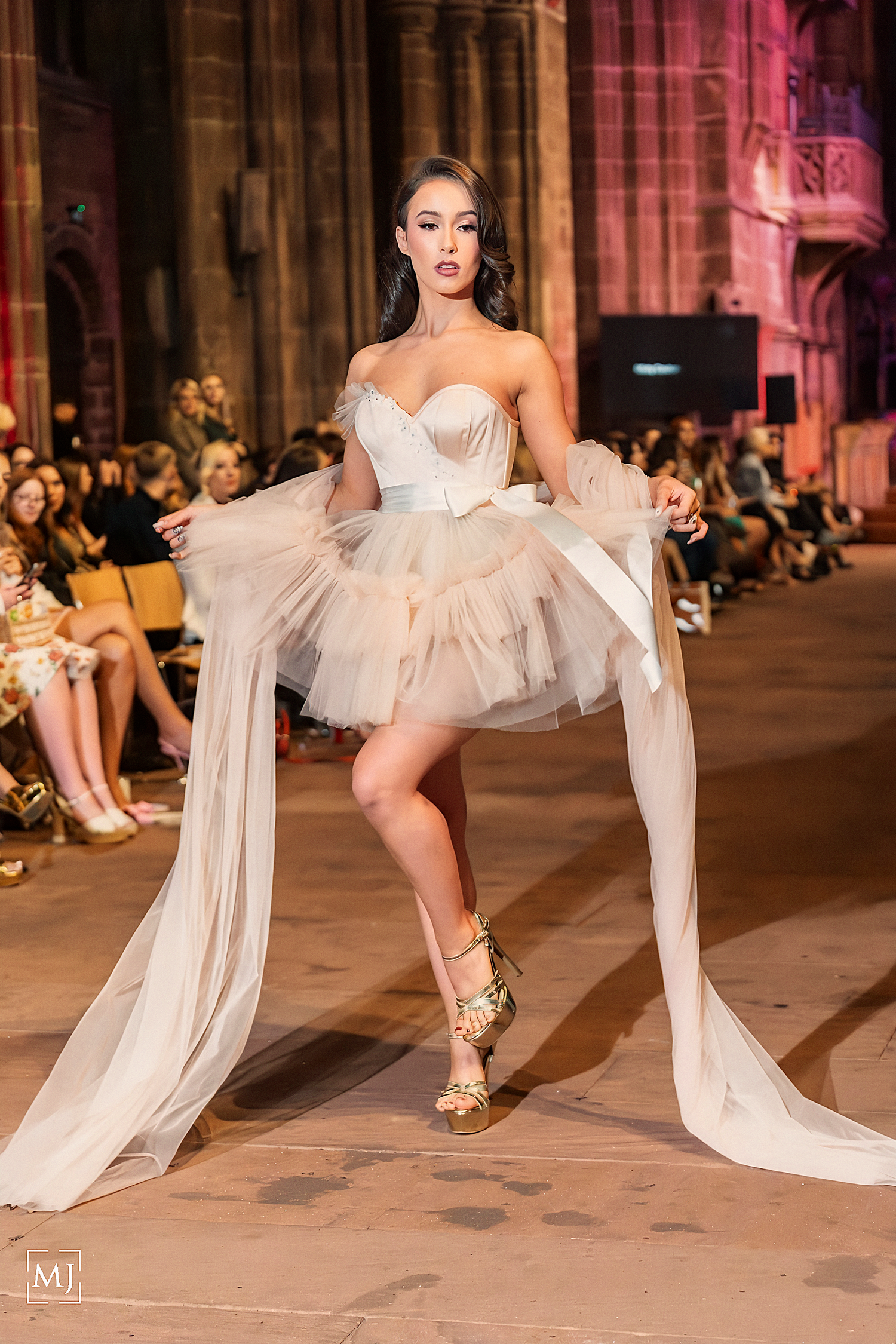 A dancer in a flowing white tulle dress performs an elegant pose during a fashion show on a warmly lit runway.
